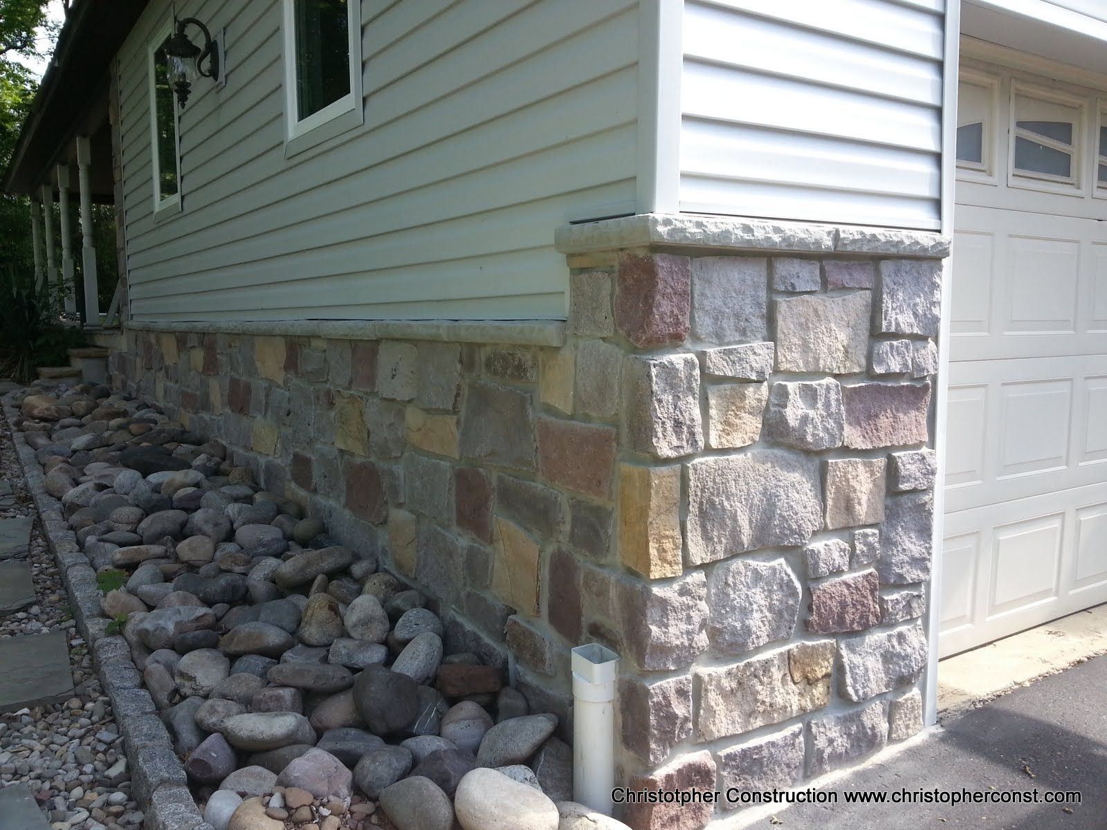 Stone-faced house corner with a garage, white siding, and a rock drainage bed along the foundation.