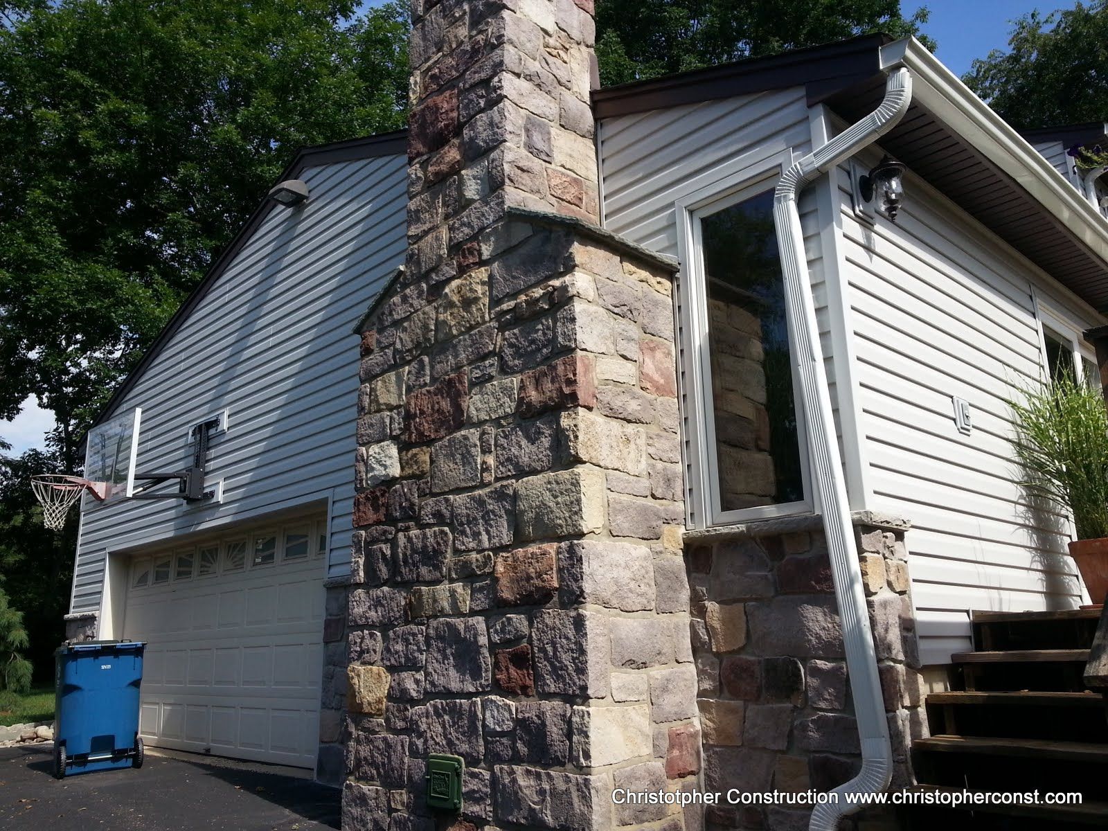 Stone chimney beside a white house with a garage and blue recycling bin