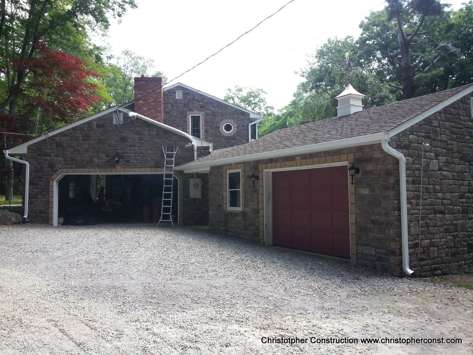 Stone house with two garage doors and gravel driveway surrounded by trees