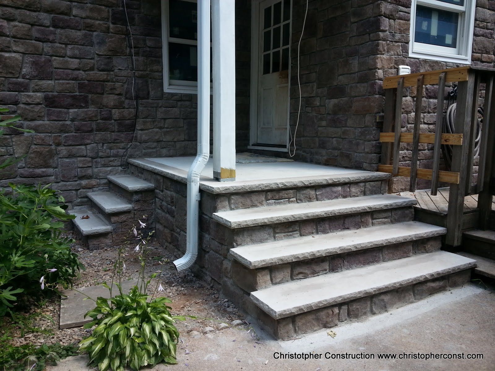 Stone house steps and porch with white downspout, beside a small garden and wooden railing.