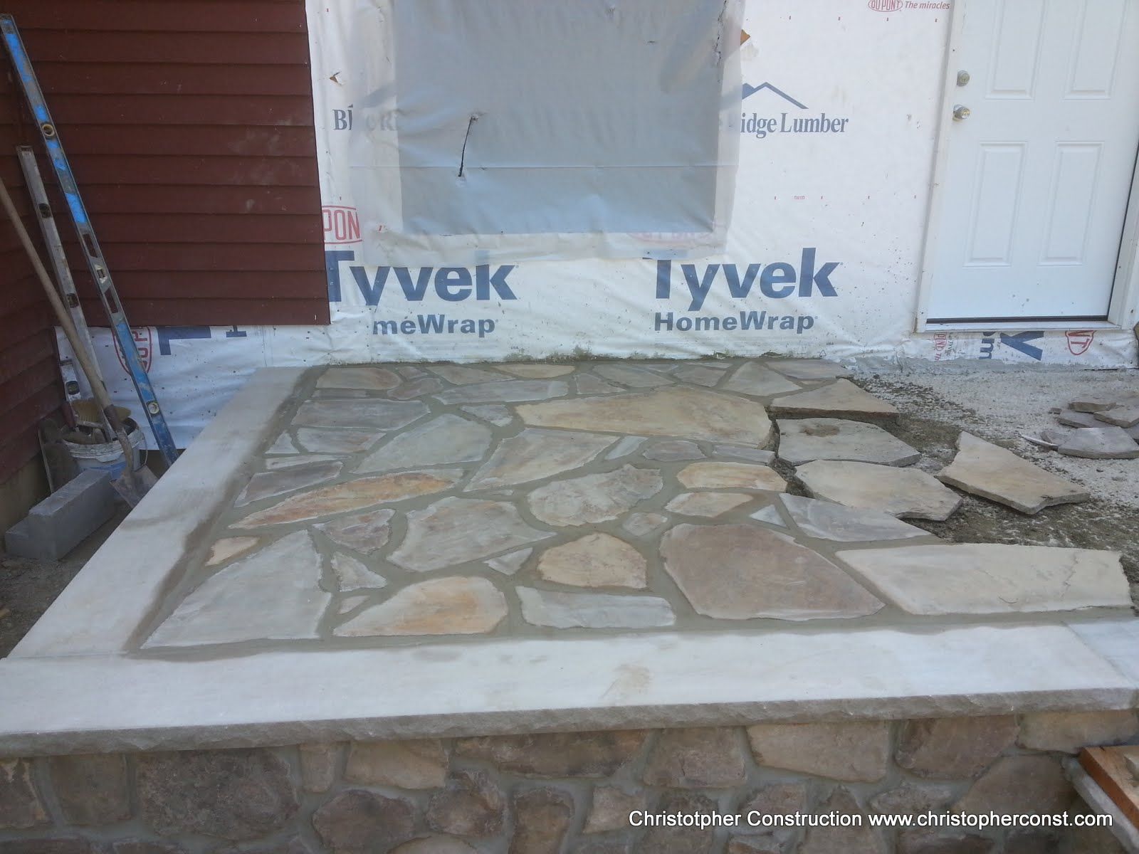 Stone patio and concrete walkway in front of a house, with Tyvek house wrap and a white door in the background