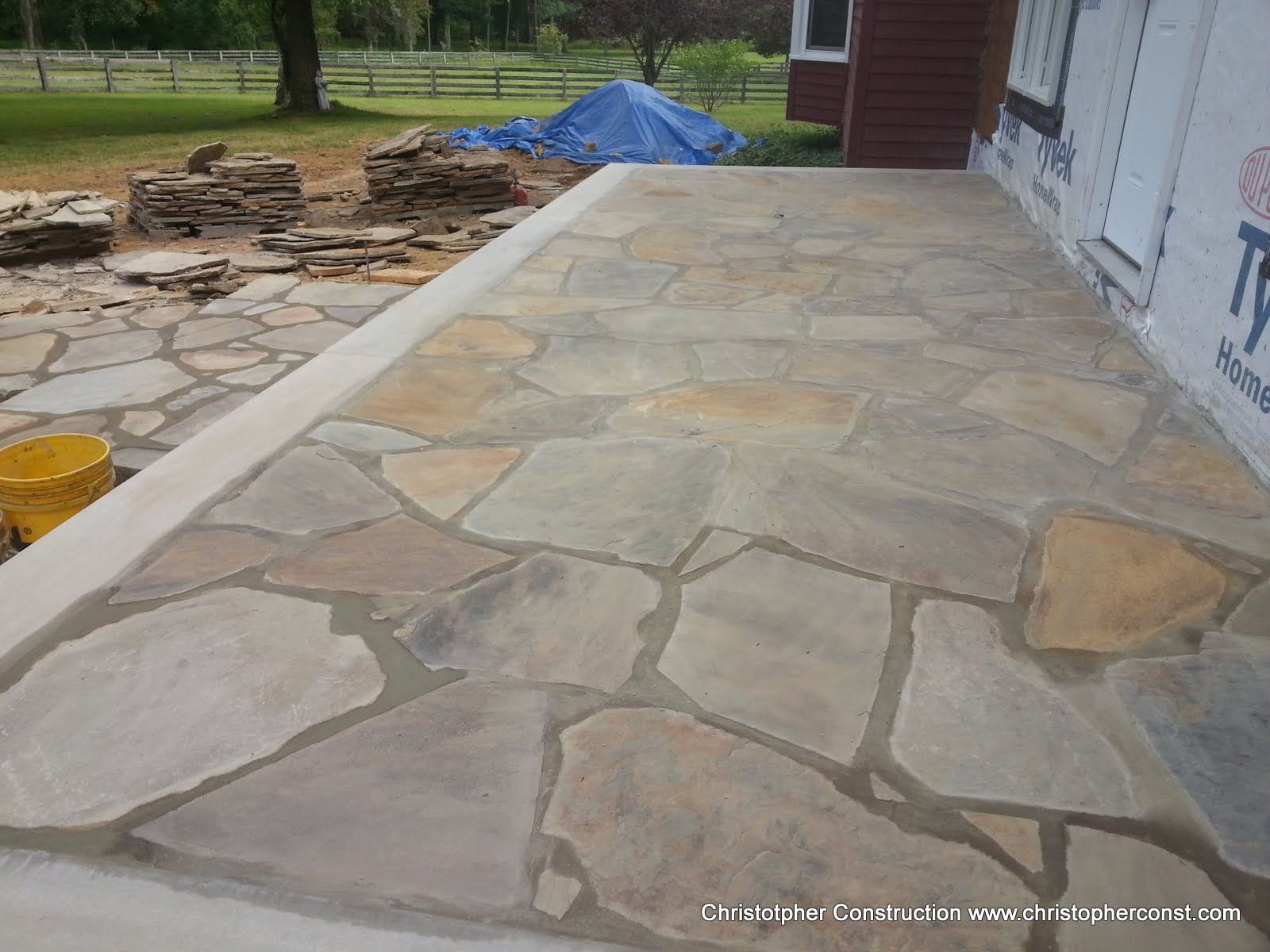 Stone patio beside a house, with stacked materials and a blue tarp in the background