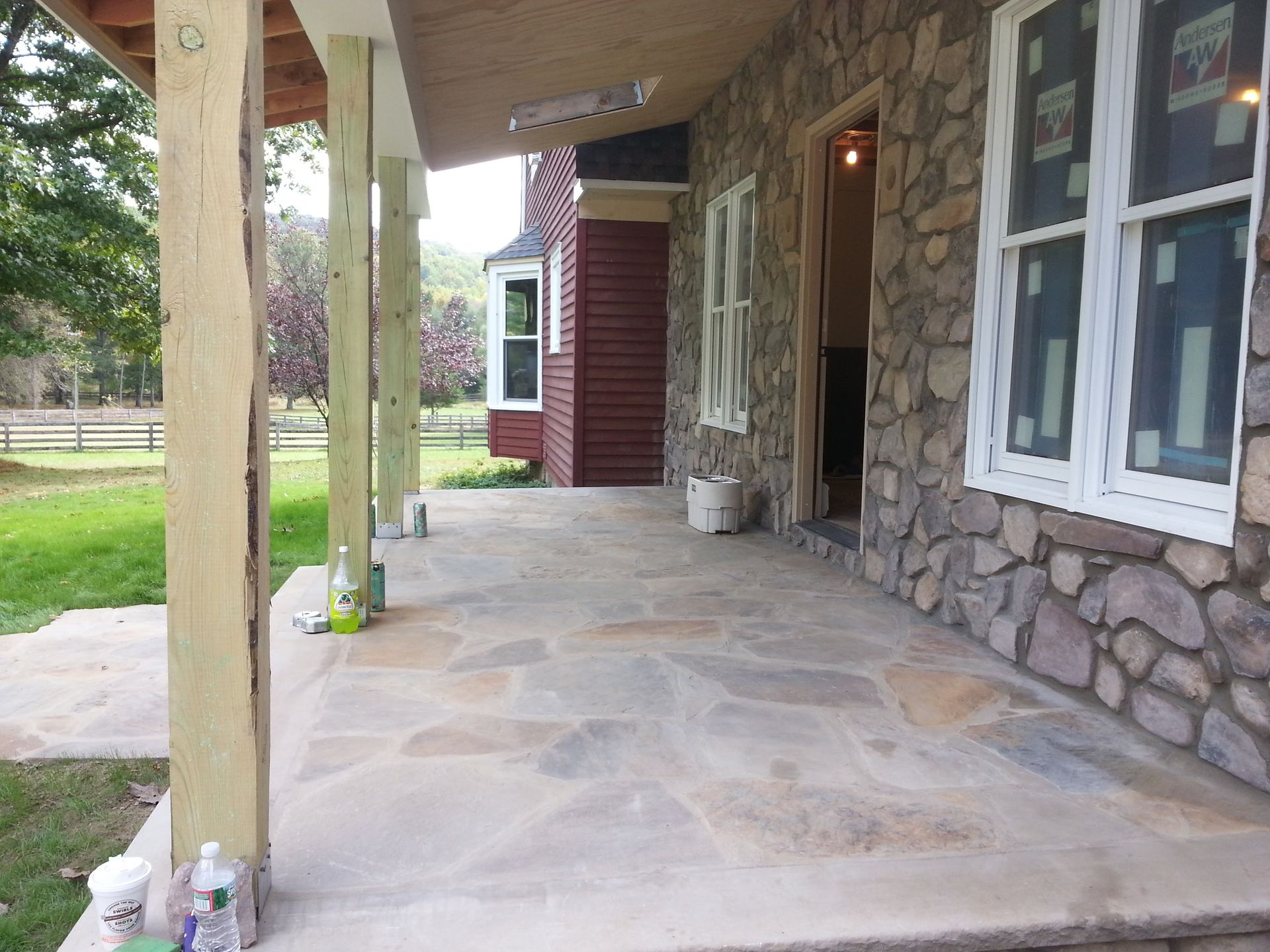 Covered stone porch with white windows, wooden columns, and a stone wall beside a grassy yard