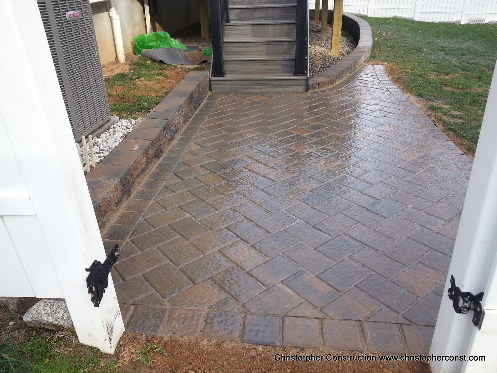 Wet brick patio walkway leading to outdoor stairs beside a white fence and house corner