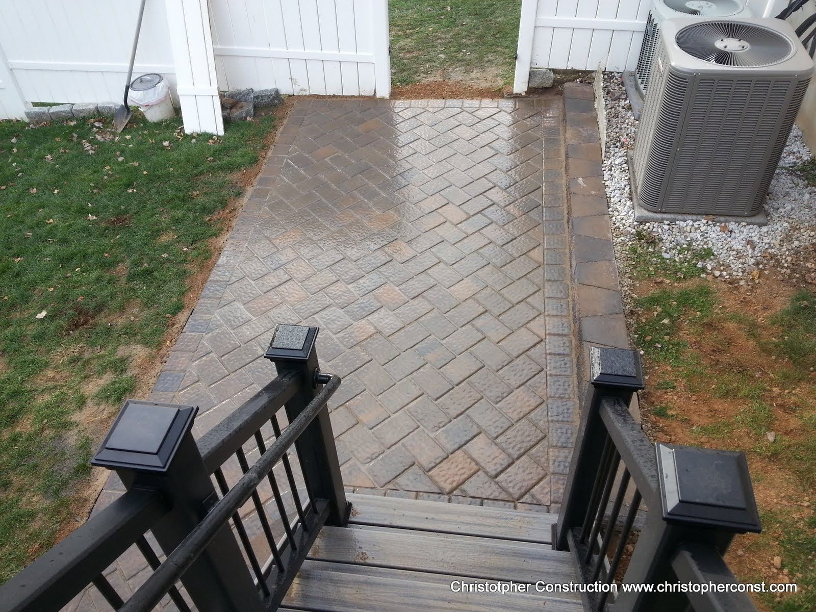 Brick walkway leading to a white fence gate, bordered by grass and a patio HVAC unit on the right.