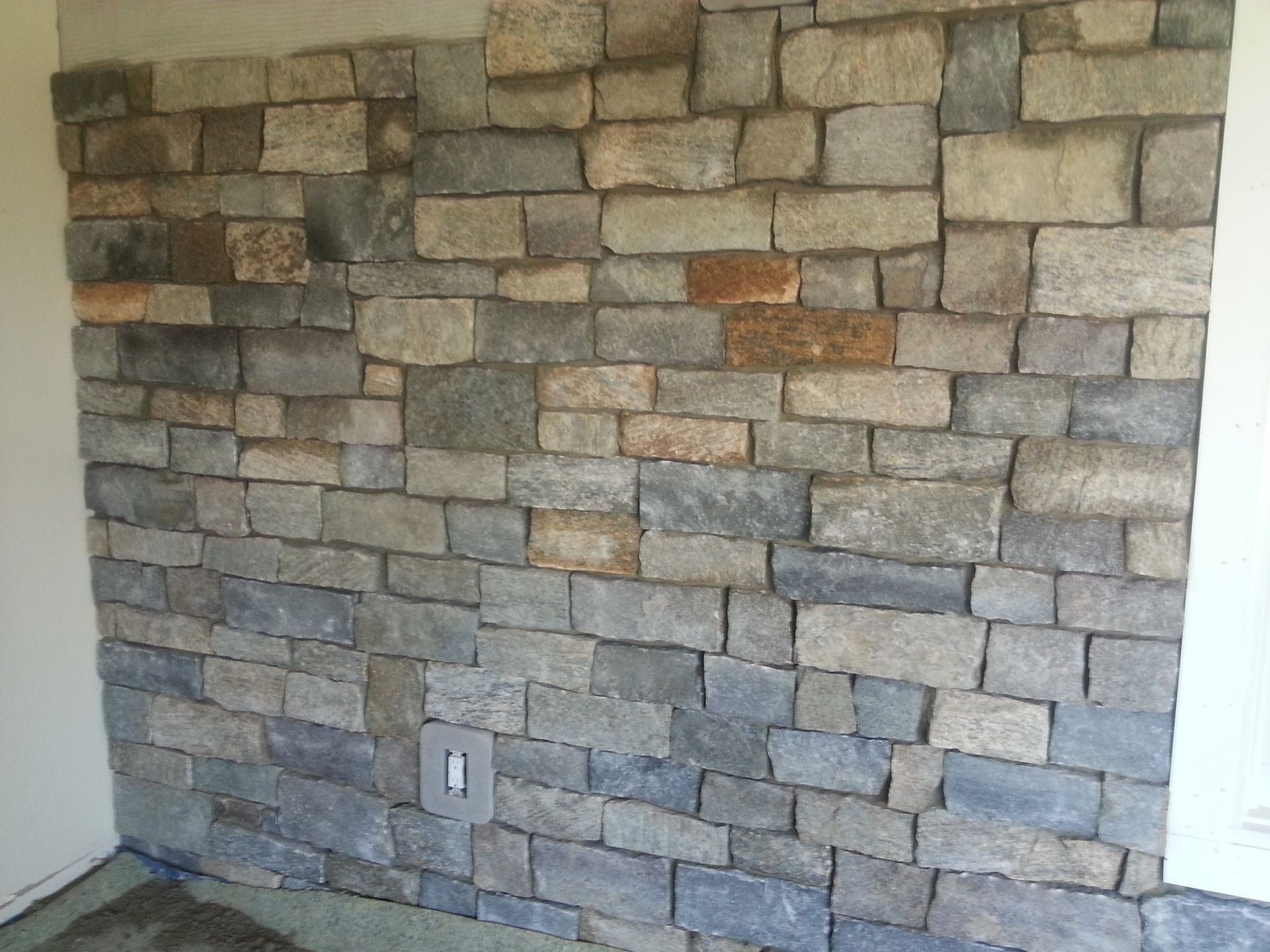 Stone veneer wall in a room corner, with gray and tan stacked stones and a visible electrical outlet.