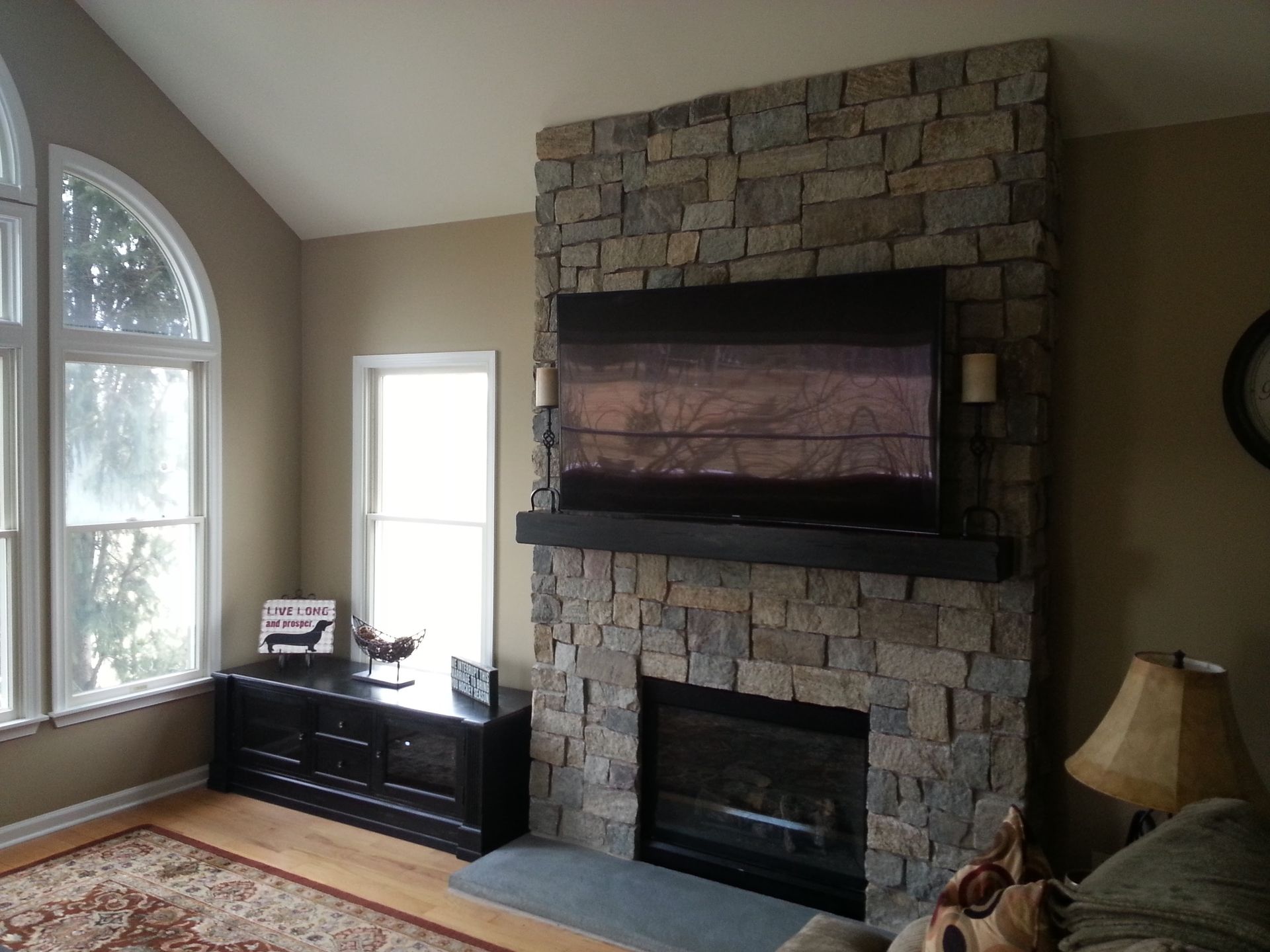 Living room with stone fireplace, wall-mounted TV, large arched window, and dark wood console