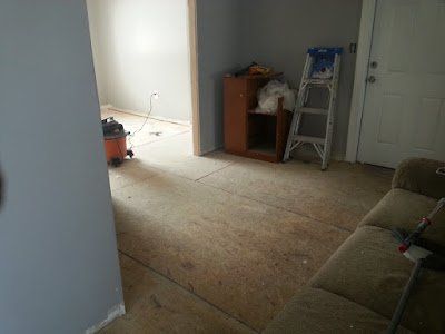 Empty room under renovation with bare carpet, ladder, small cabinet, and sofa at right.