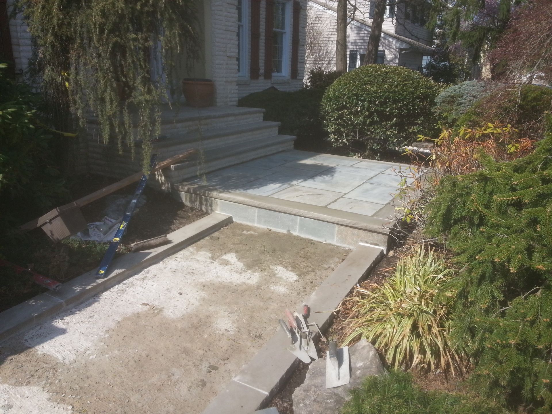 Front walkway with steps and a sloped concrete ramp beside a house, surrounded by shrubs and plants.