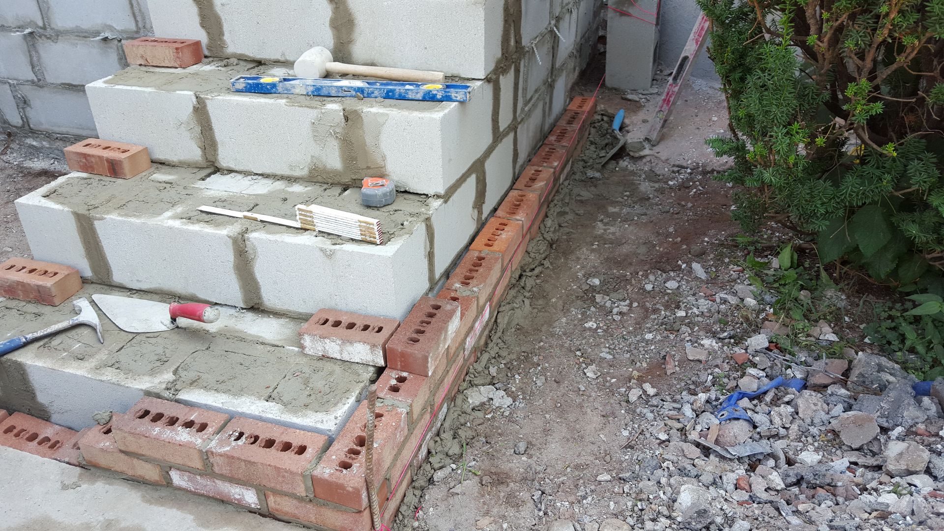 Partially built brick wall beside a narrow dirt path, with rubble and greenery on the right.
