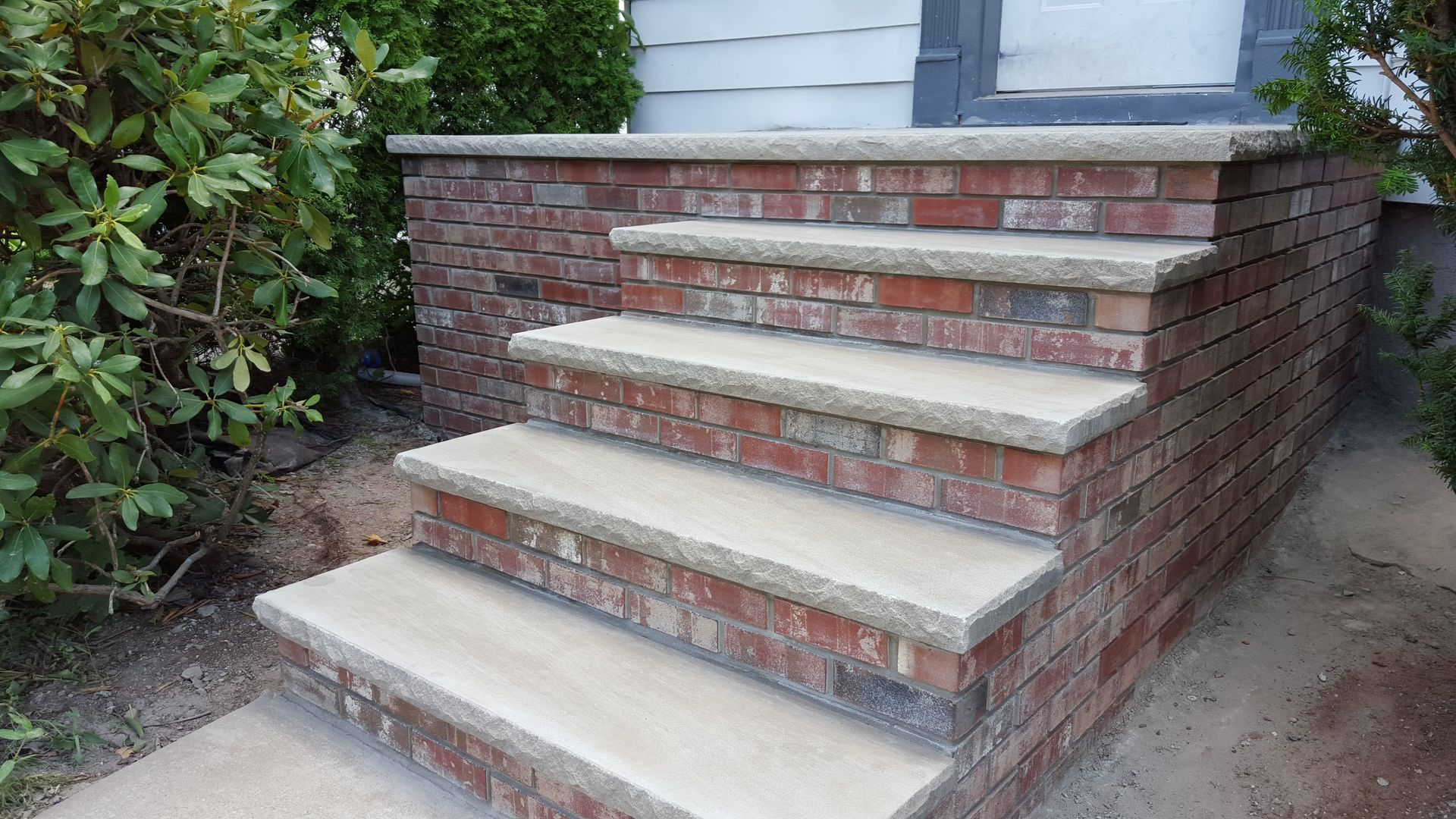 Brick front steps leading to a house porch with white trim and green shrubs nearby