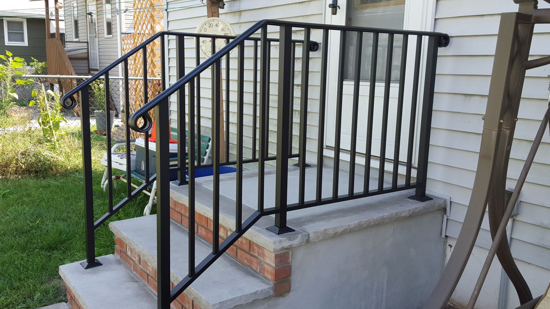 Black metal railing on concrete front steps beside a house with light siding and a small garden.