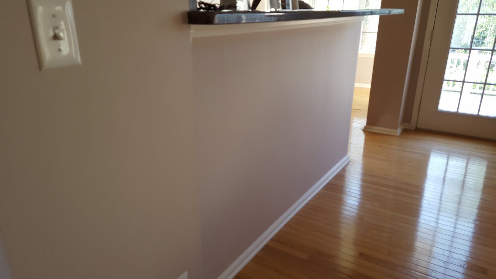 Empty hallway with light wood floors, beige walls, and a small breakfast bar near a bright glass door.