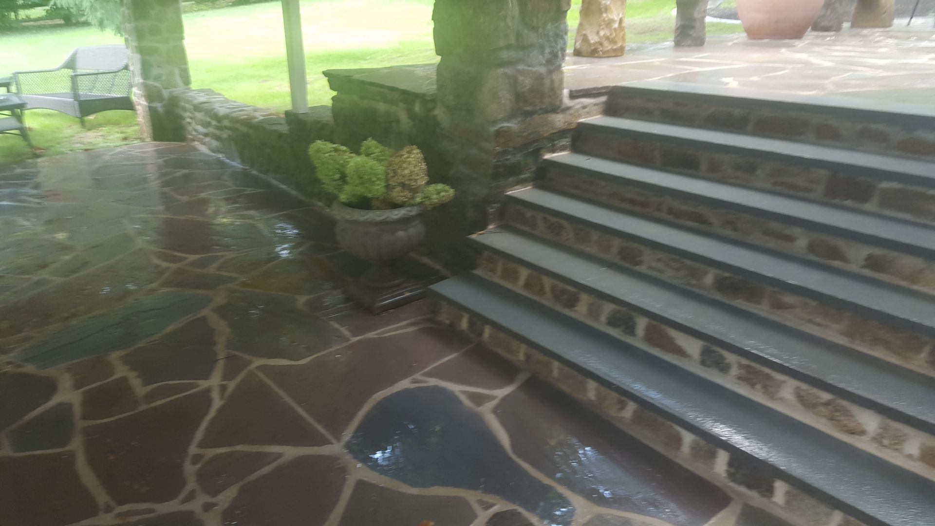 Wet stone patio beside gray steps and potted plants under a shaded porch
