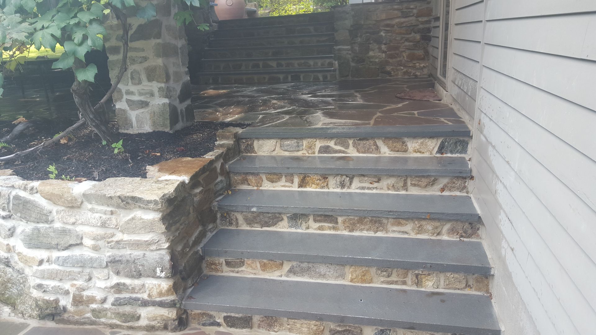 Stone steps leading to a raised walkway beside a white-sided house and stone retaining wall