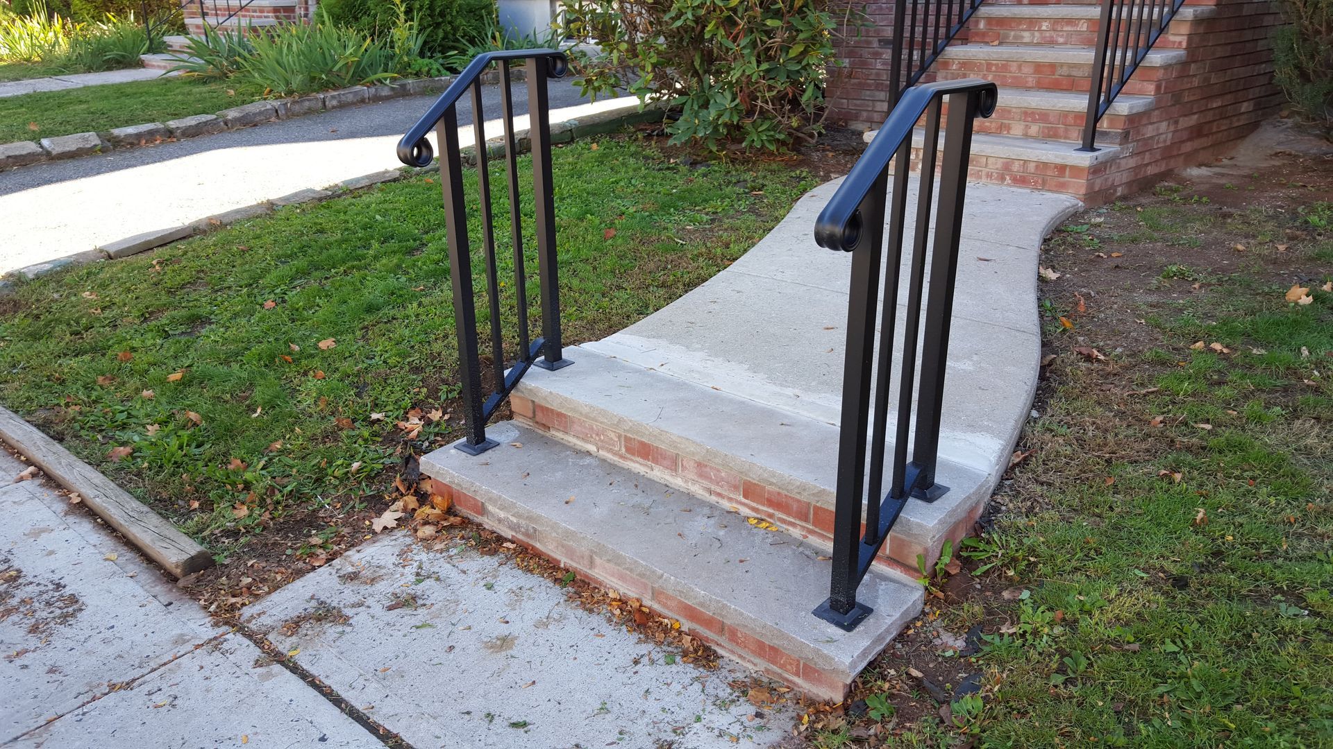 Concrete walkway with steps and black handrails beside a grassy slope and brick path