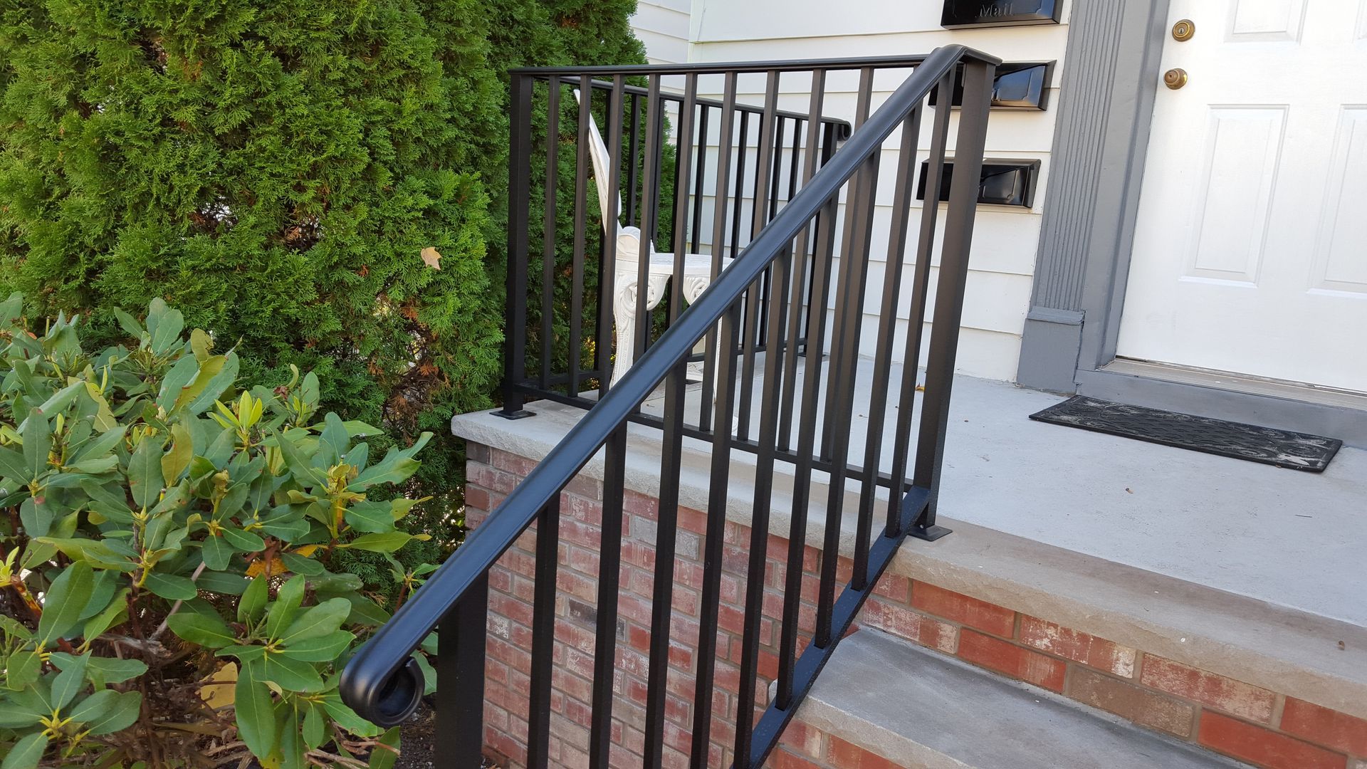 Black metal handrail beside brick steps and a white front door by a trimmed hedge.