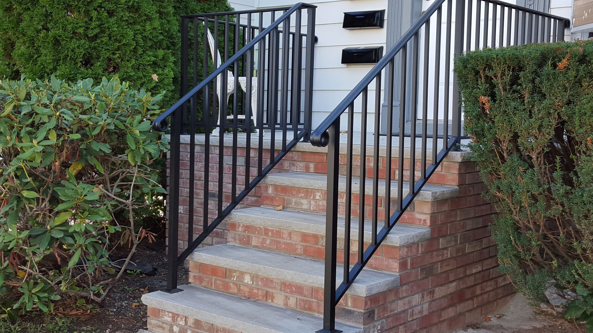 Brick front steps with black metal handrails leading to a doorway, flanked by trimmed hedges