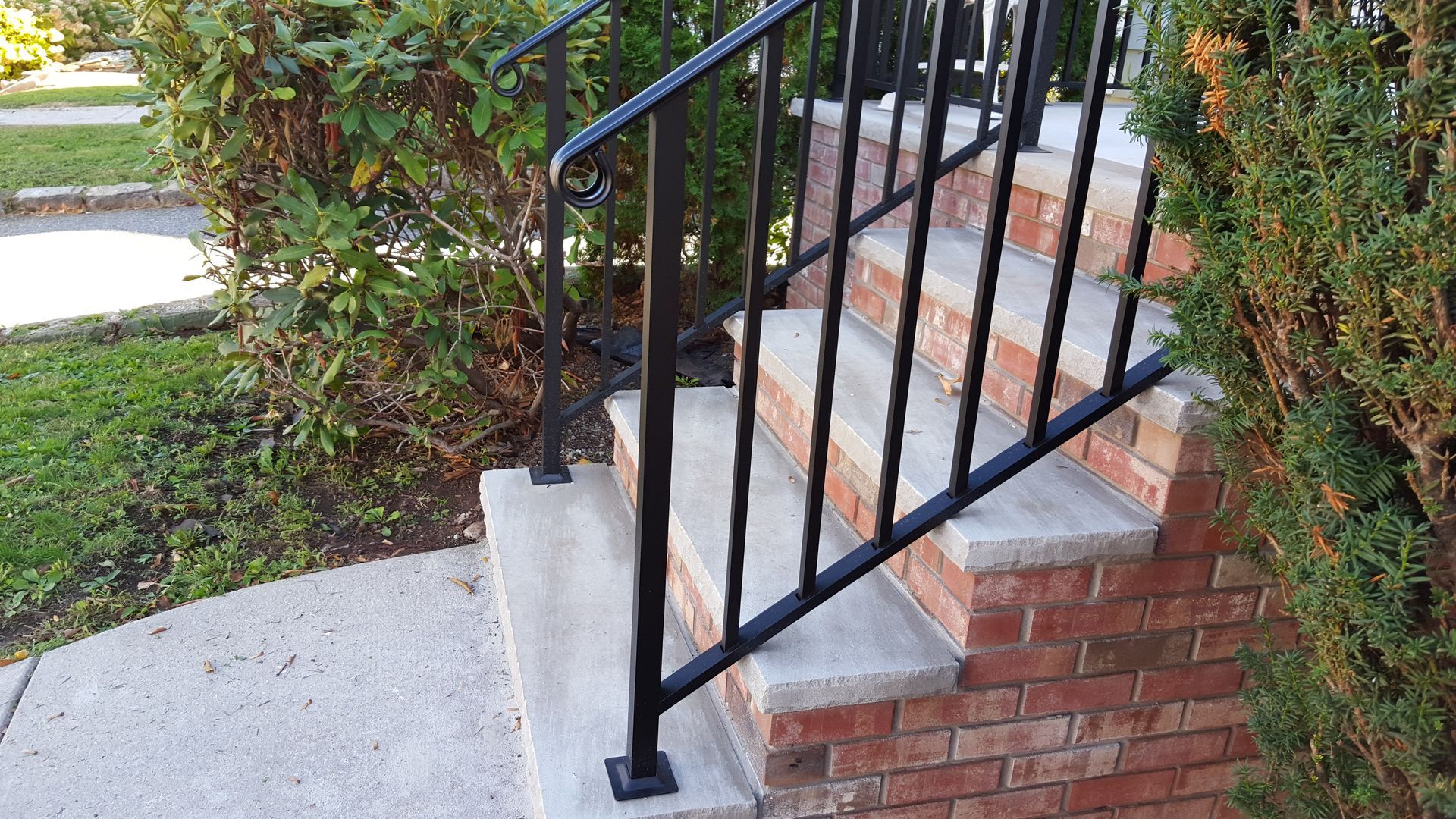 Brick front steps with black metal railing beside a concrete walkway and shrubs