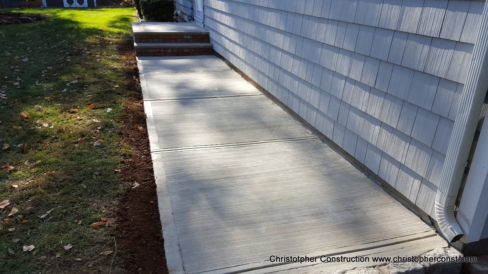 Concrete walkway beside a white-sided house, with sunlight and shadows along the path.