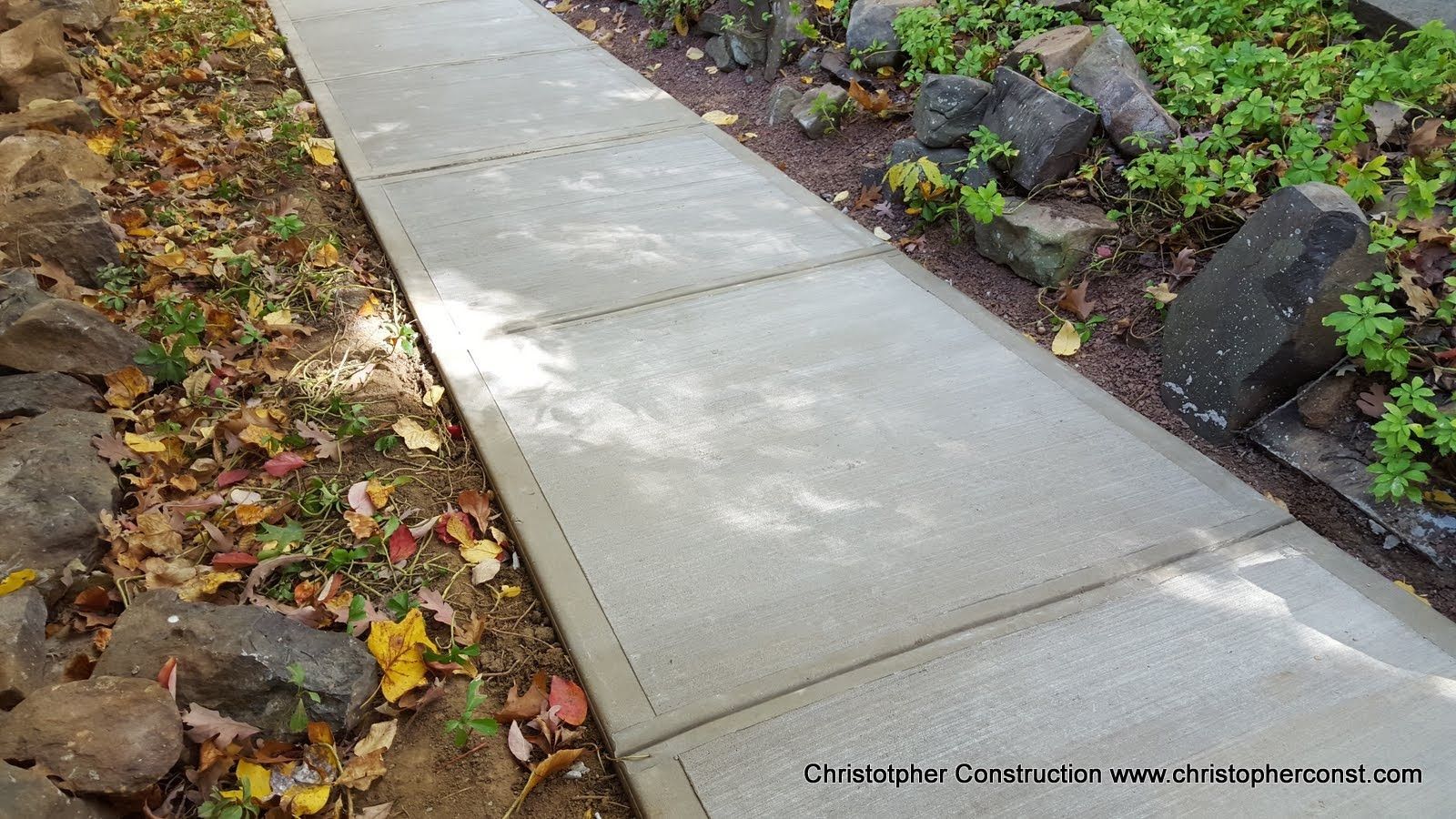 Concrete sidewalk bordered by fallen leaves and a landscaped garden bed with flowers and mulch