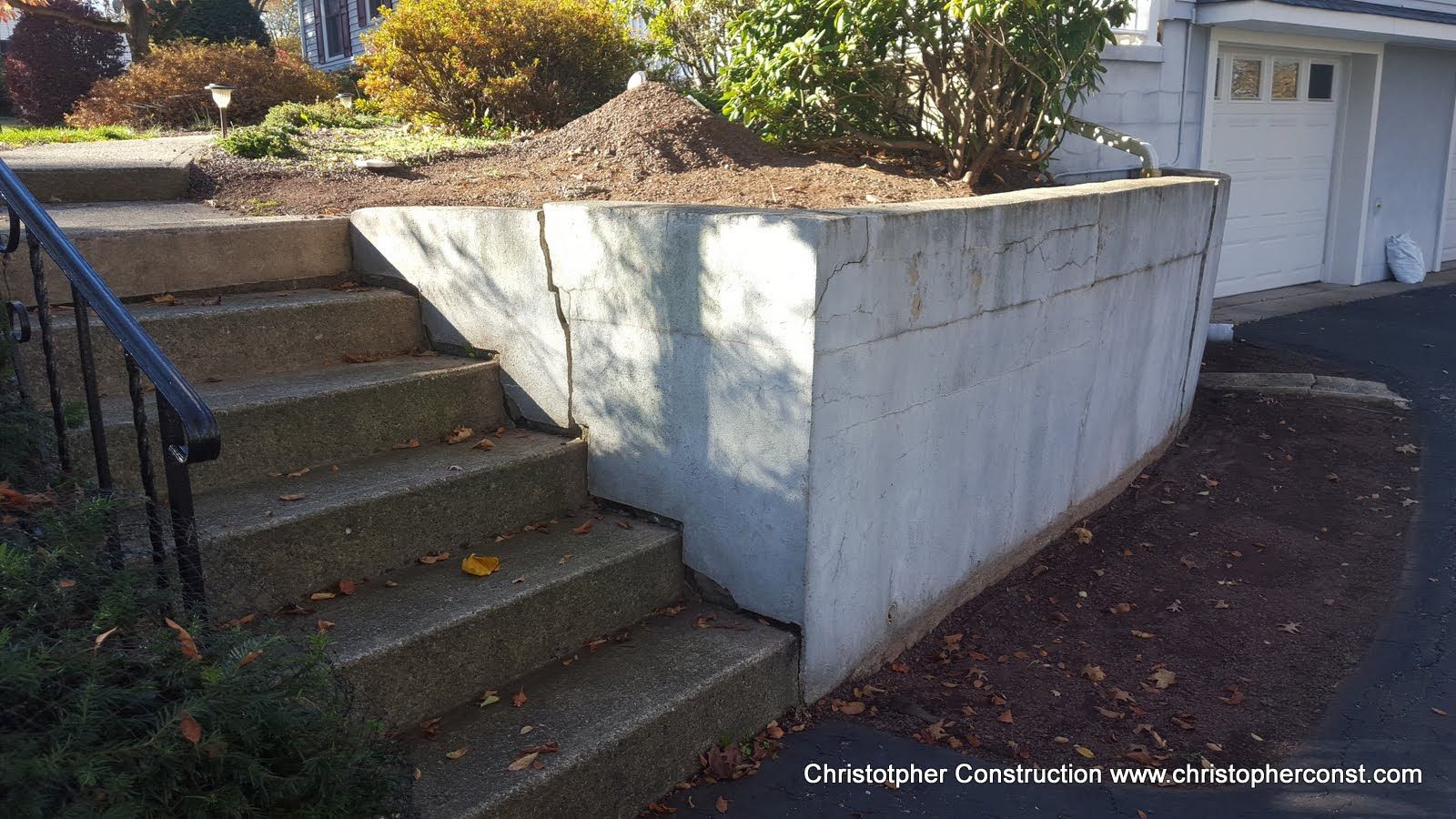 Concrete retaining wall beside outdoor steps and a driveway, with shrubs on top.