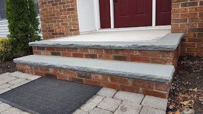 Brick front steps with concrete treads leading to a red door, beside a paved walkway.
