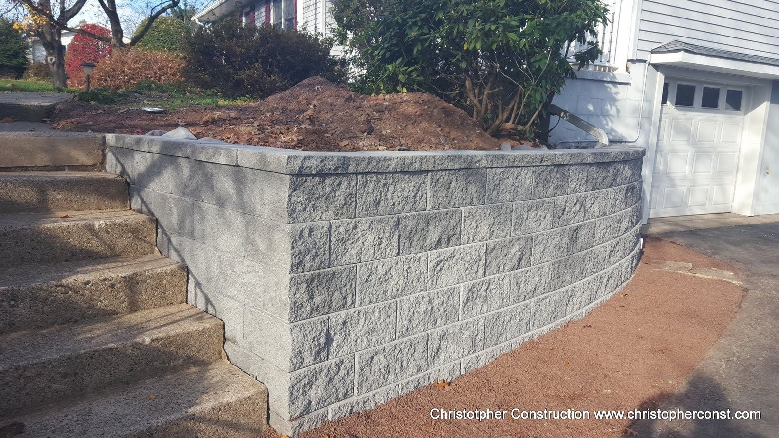 Gray concrete retaining wall beside stone steps and driveway, with landscaped soil and a white garage door.