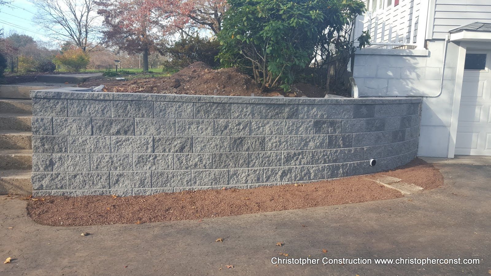Curved gray stone retaining wall beside a white gate and trees in a landscaped outdoor area