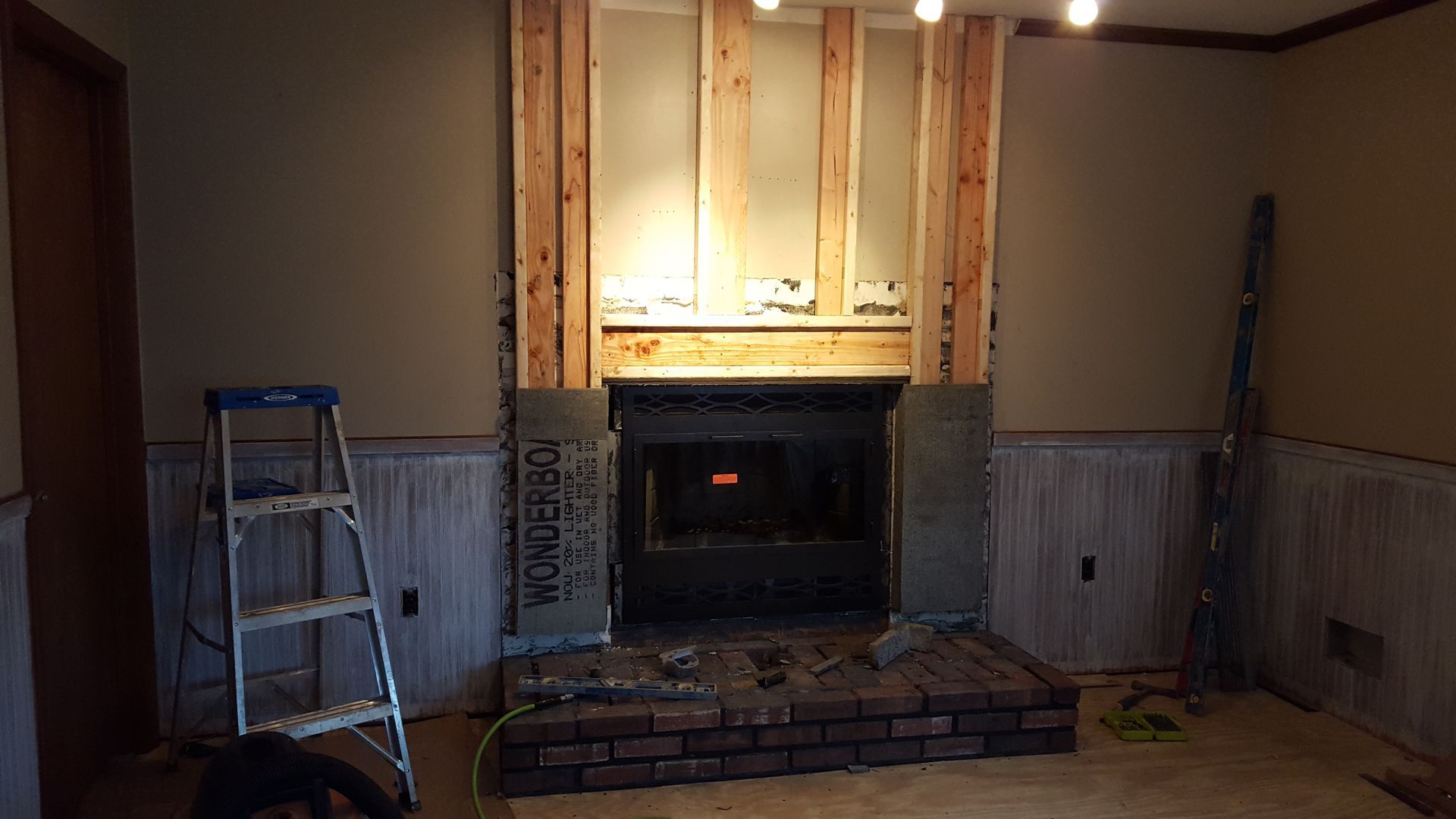 Living room with a fireplace under renovation, ladder on the left and exposed wall framing around the mantle