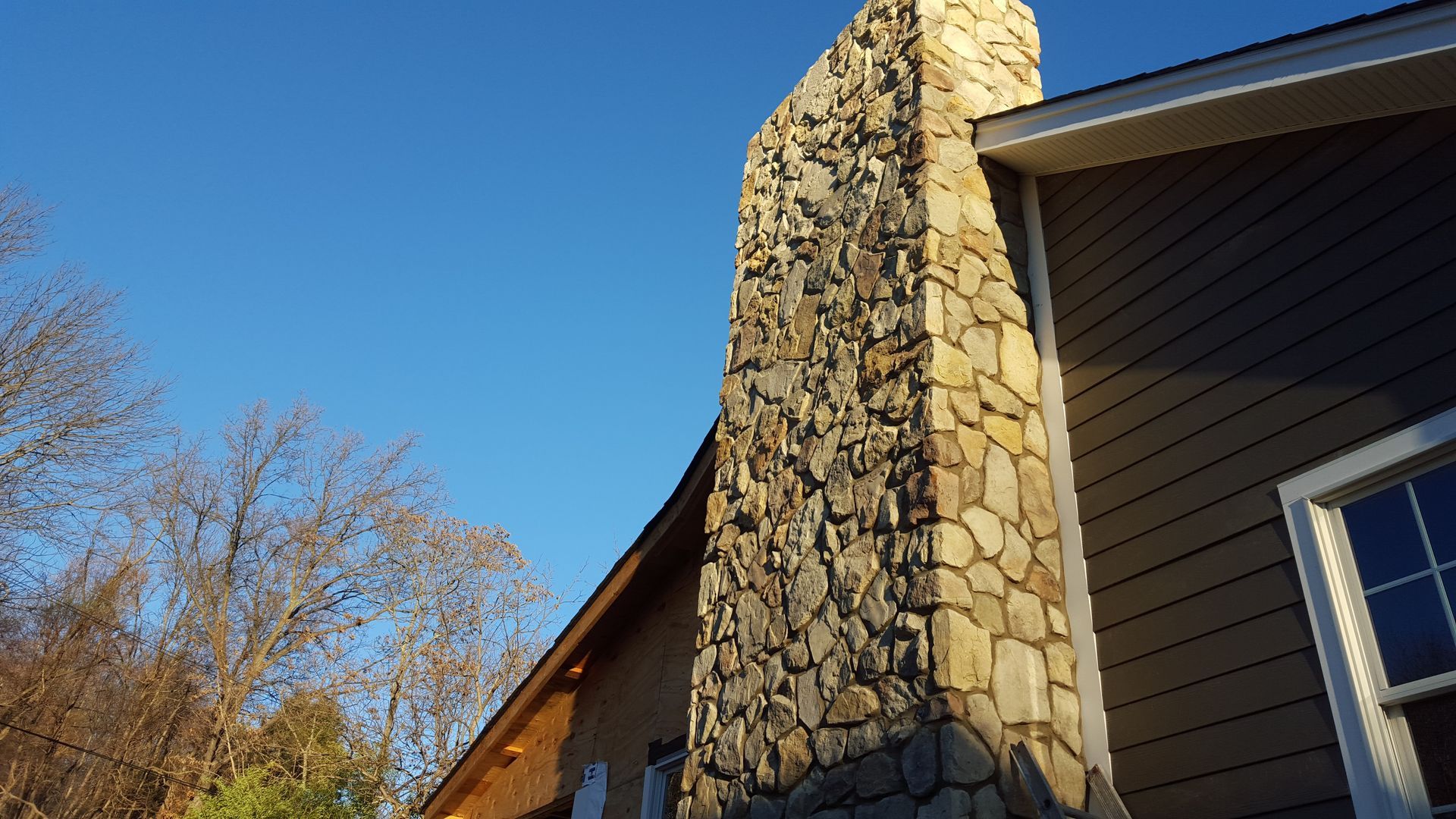 Stone chimney beside a dark house under a clear blue sky, with bare trees in the background