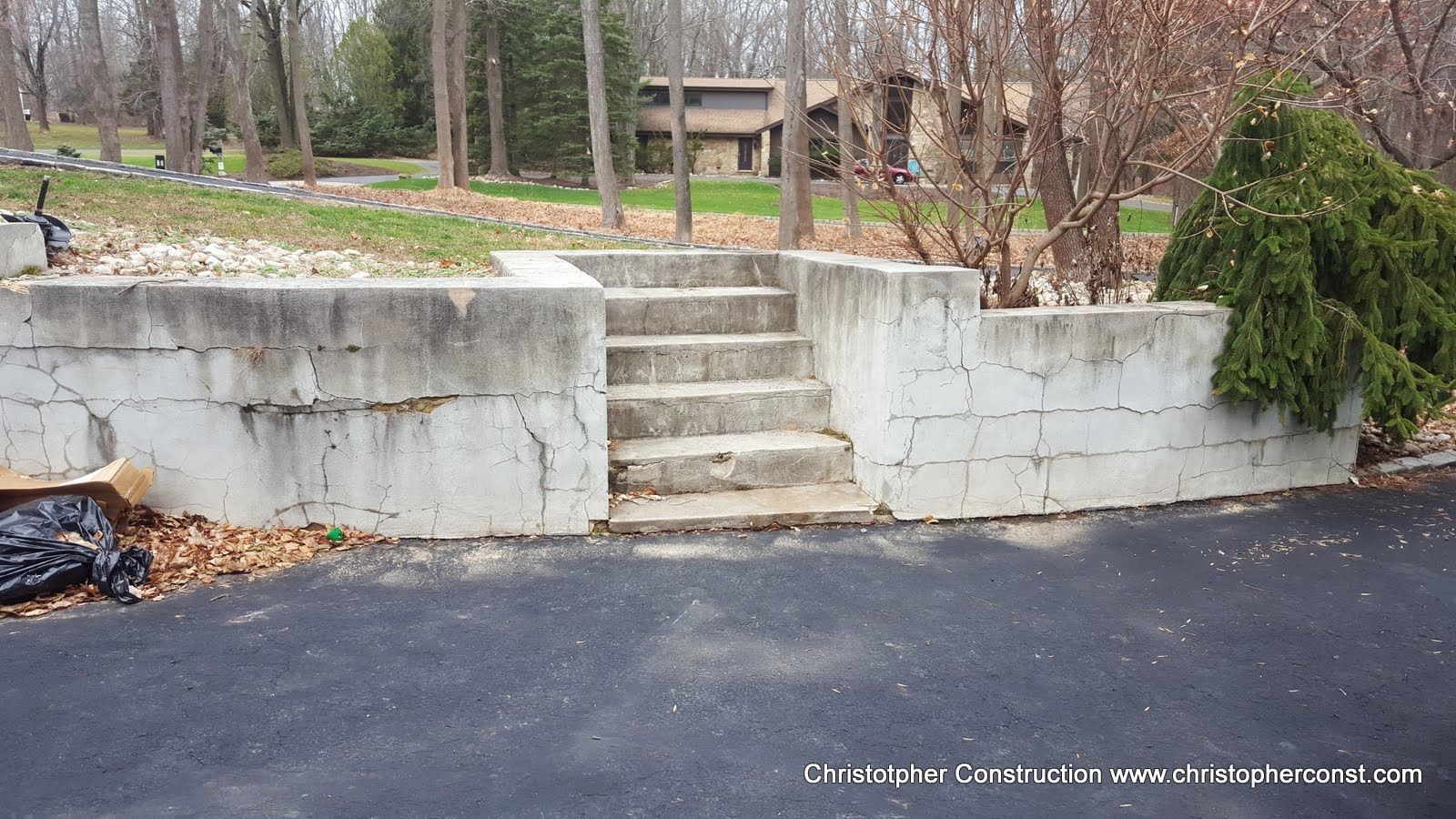 Concrete retaining wall with center steps leading to a grassy yard and trees beside an asphalt driveway.