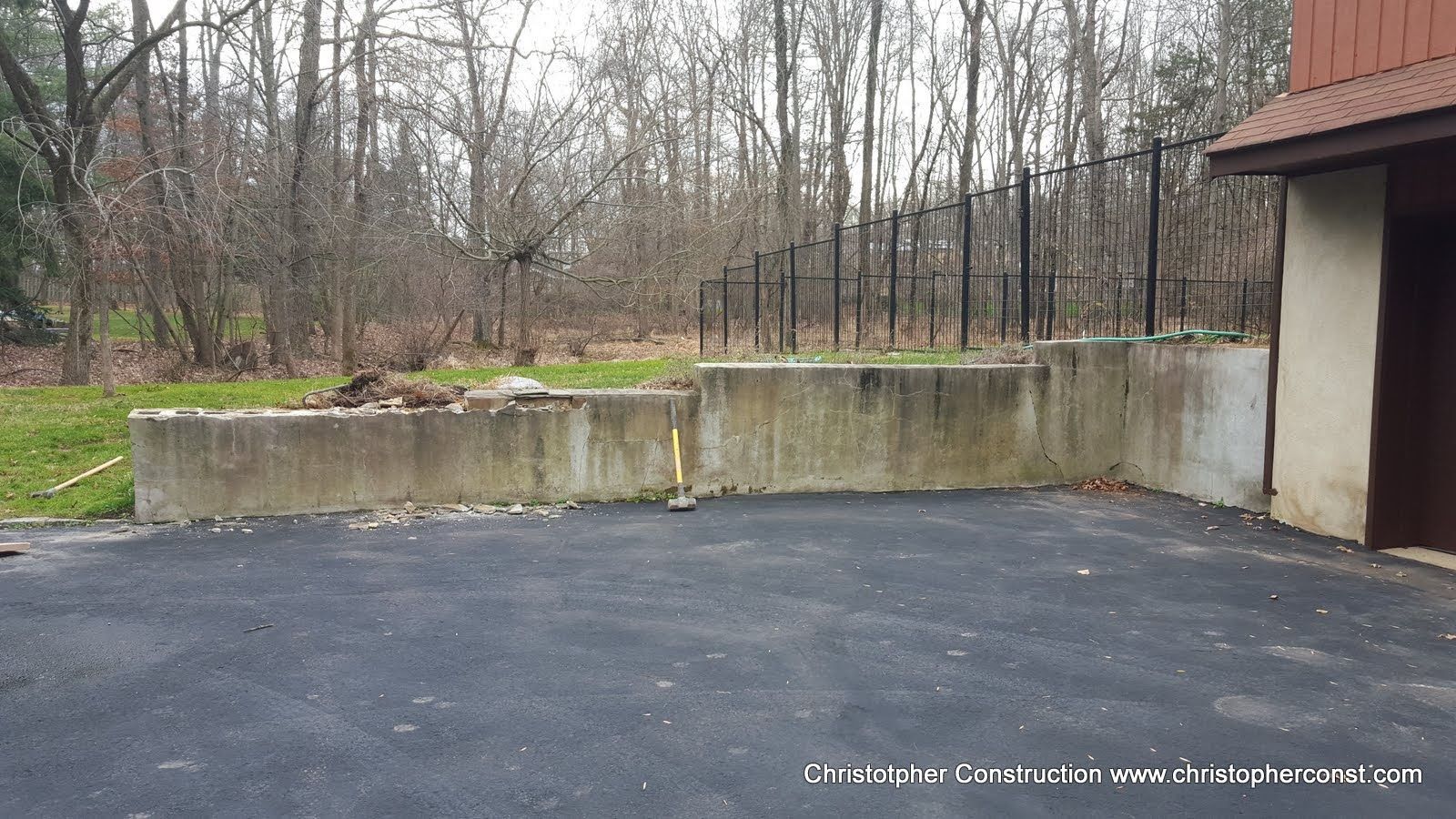 Empty asphalt driveway beside a concrete wall and garage, with leafless trees in the background.