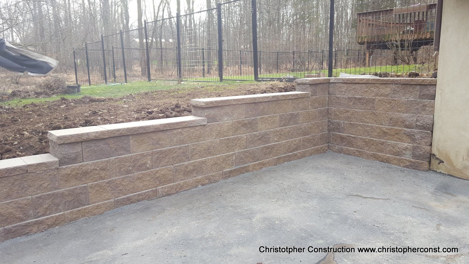 Empty concrete patio with a low tan retaining wall beside a wooded yard