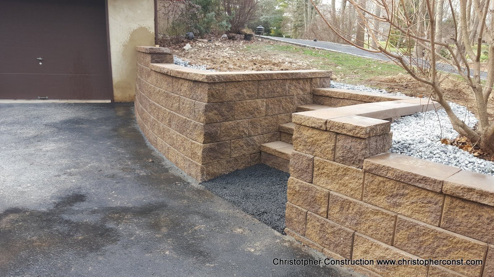 Stone retaining walls beside a driveway and garage, with light snow on the ground and shrubs above