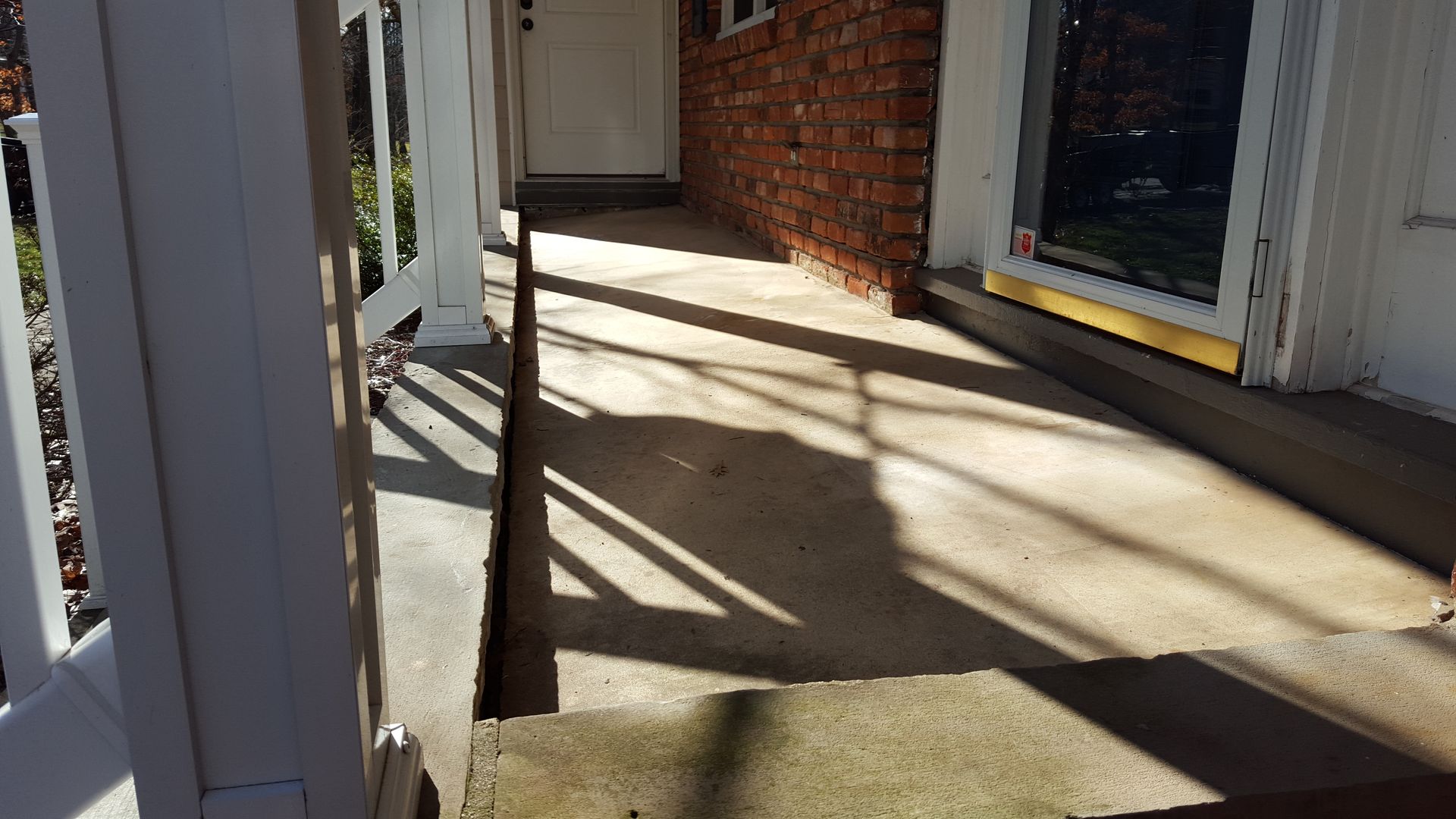 Sunlit porch walkway with white railings, brick wall, and strong window shadows on concrete floor
