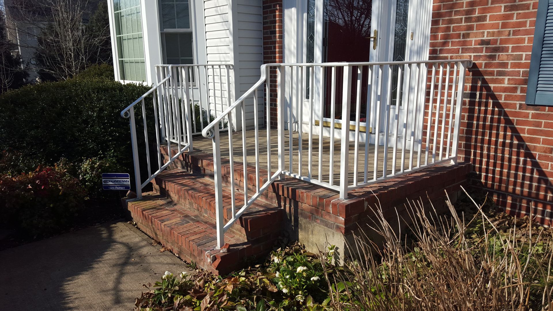 Brick front porch with white railing, steps, and a red front door beside bushes