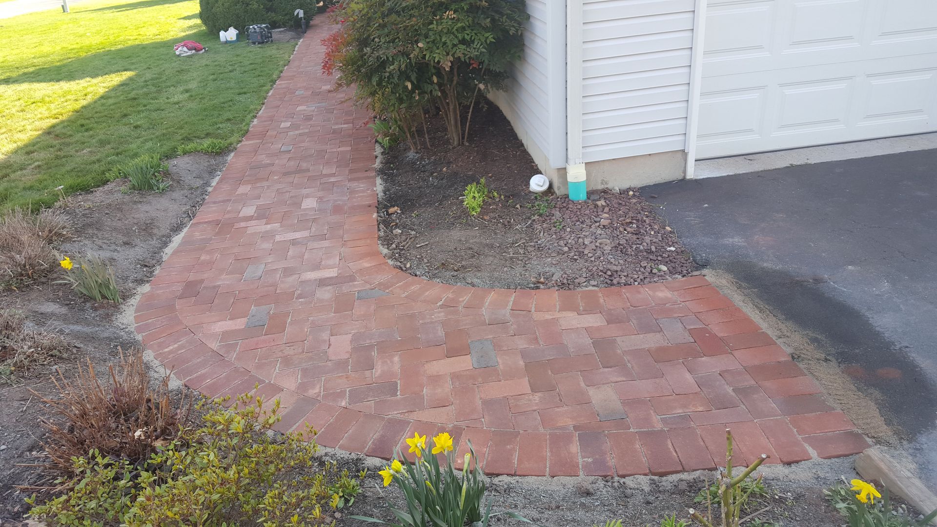 Brick garden path curving beside a white house and driveway, bordered by mulch and yellow flowers