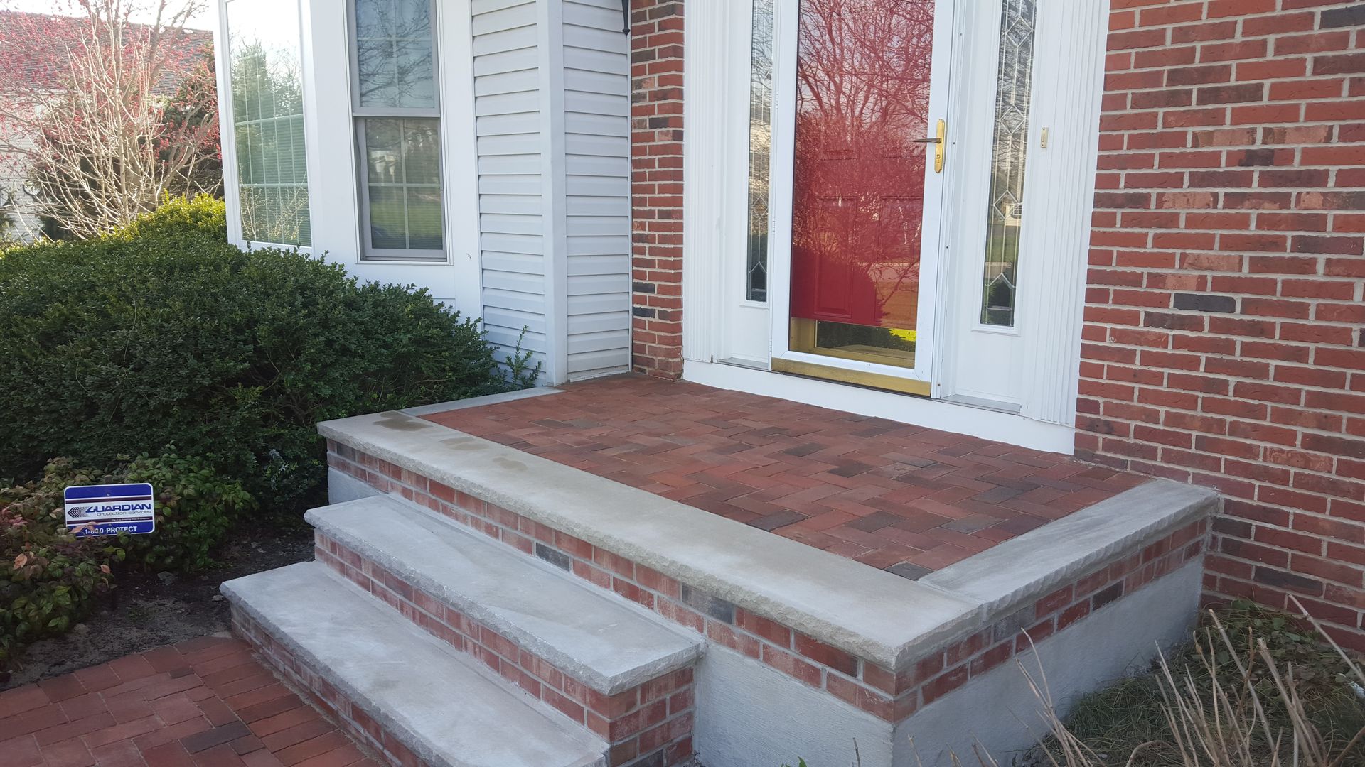 Brick front porch with concrete steps leading to a red front door and white trim on a house