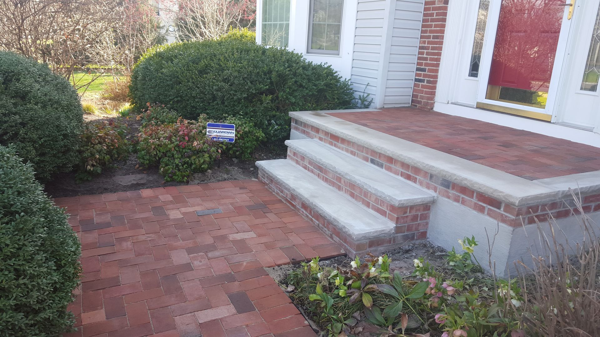 Brick front porch with two steps, red door, and landscaped shrubs along a walkway.