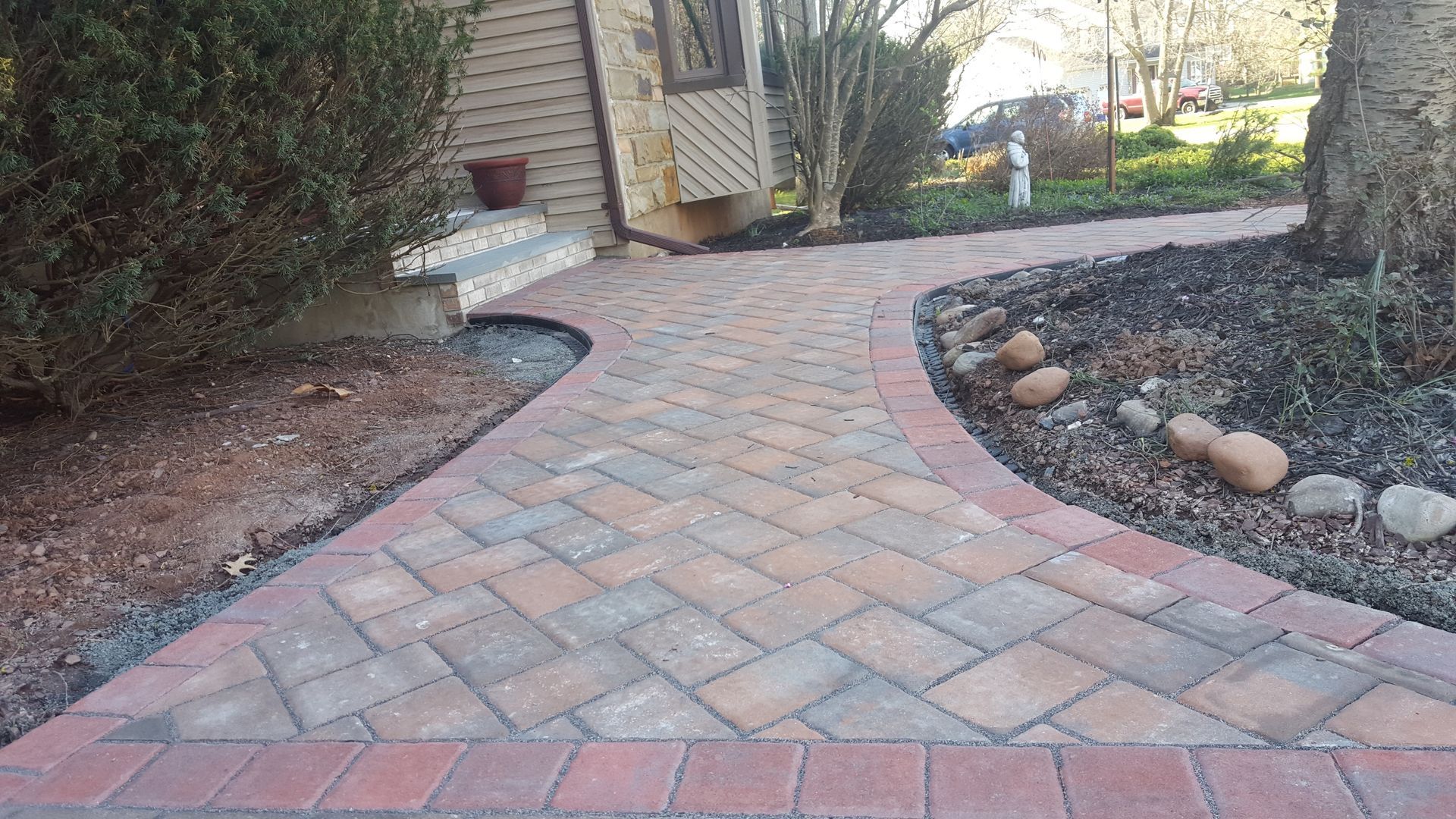 Curved brick garden pathway bordered by rocks and shrubs leading to a house entrance