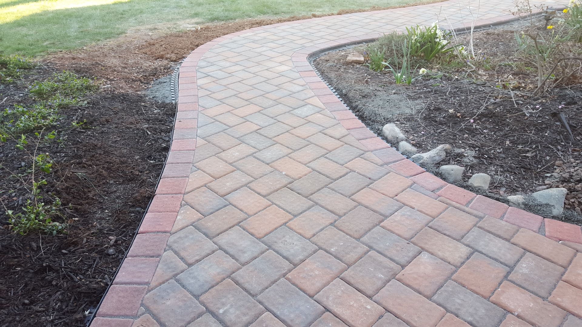 Curved brick paver walkway bordered by soil and rocks in a landscaped garden area