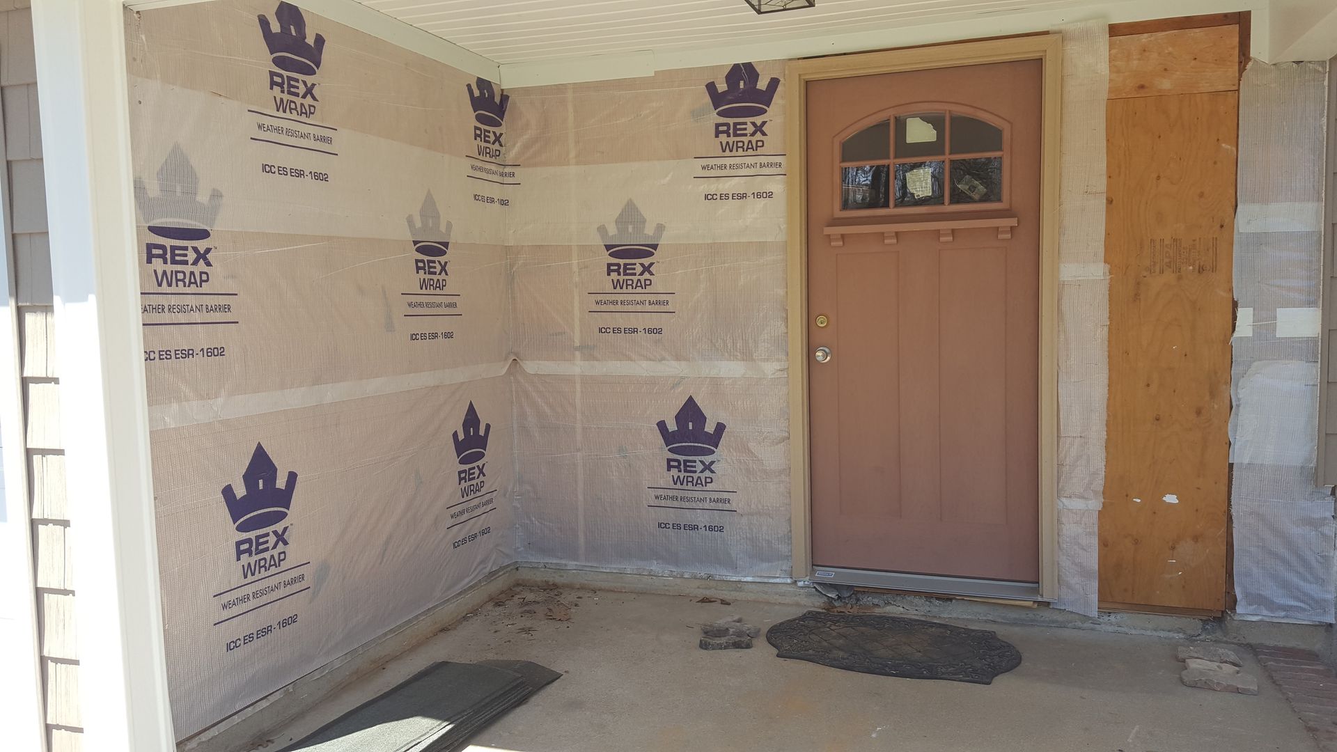 Unfinished front porch with wrapped walls, a tan door, and construction materials on the floor.