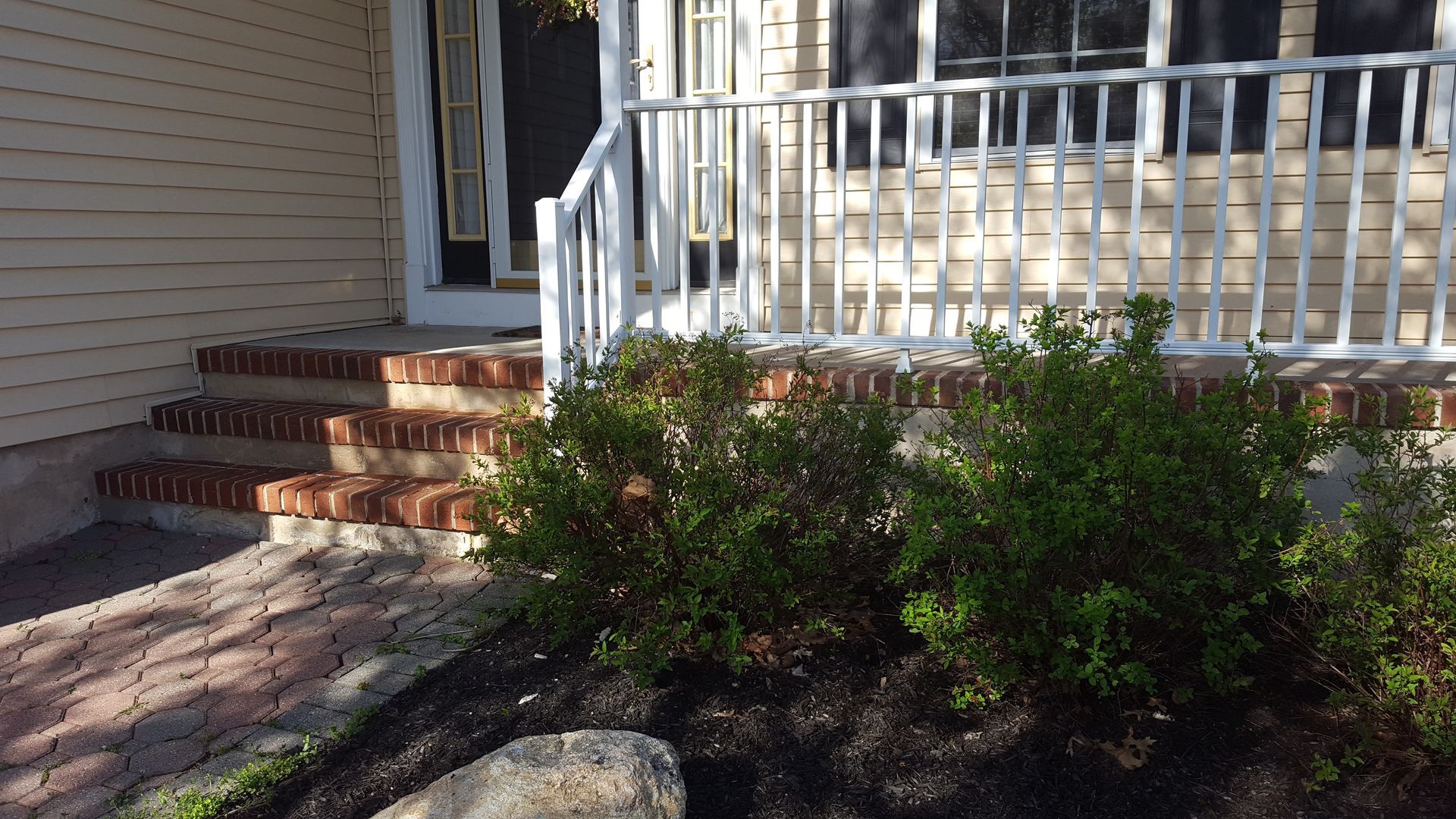 Front porch with white railing, brick steps, and shrubs beside a house entrance