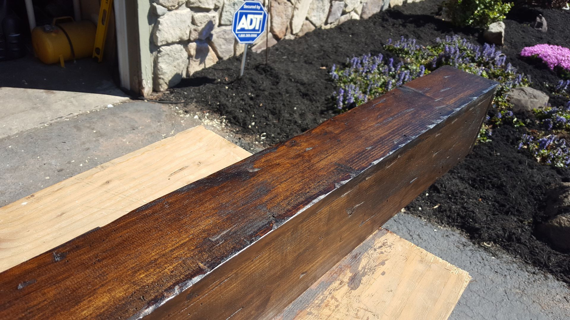 A long, polished wooden beam on sawhorses beside a stone house and dark mulch garden bed.