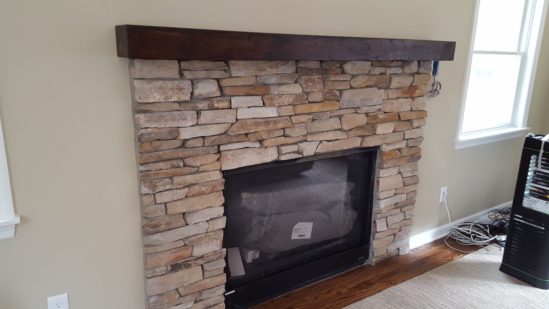 Stone fireplace with dark wood mantel in a bright room, with a window and hardwood floor nearby
