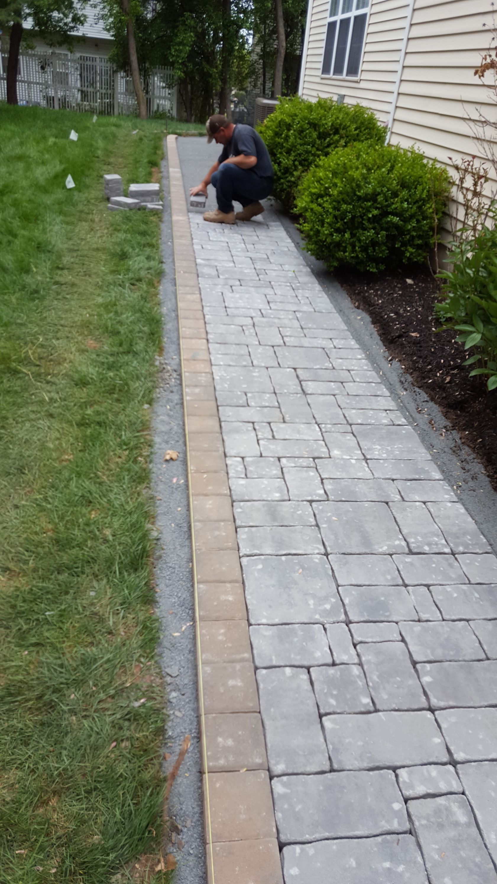 Person kneeling beside a newly laid gray paver walkway next to a house and lawn