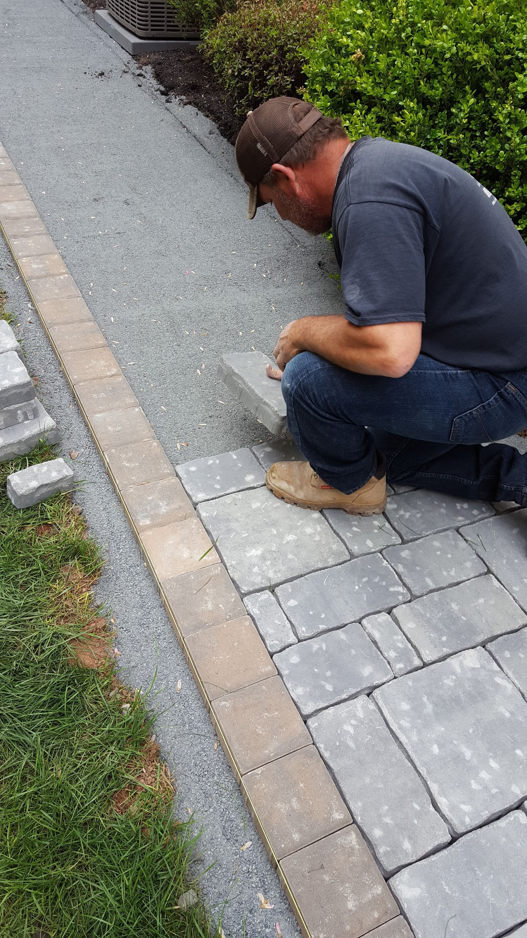 Man kneeling on a paved walkway, inspecting or repairing a brick edge beside gravel and grass.