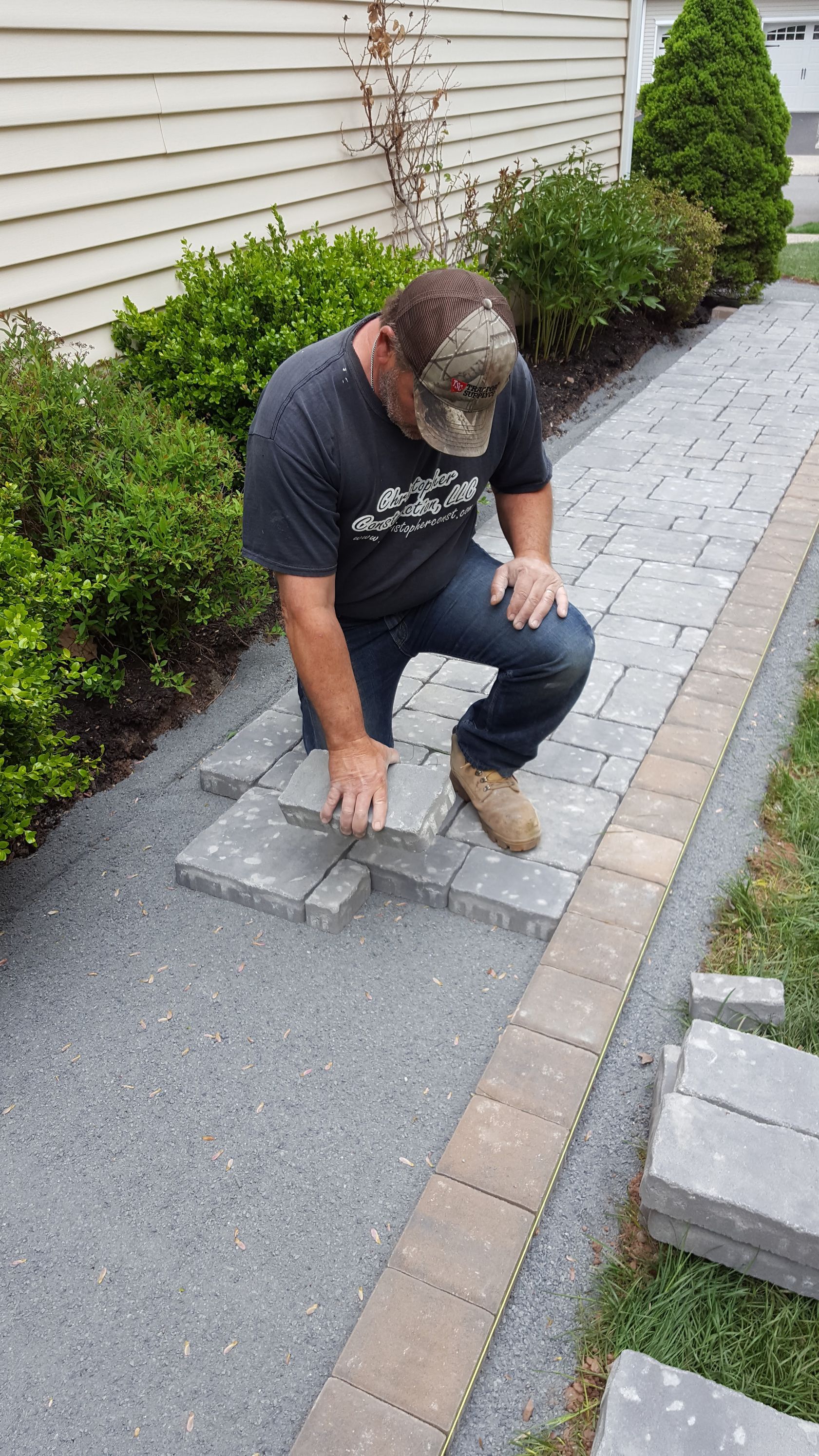 Person kneeling on a driveway, pointing at a cracked patch in the pavement beside a landscaped yard.