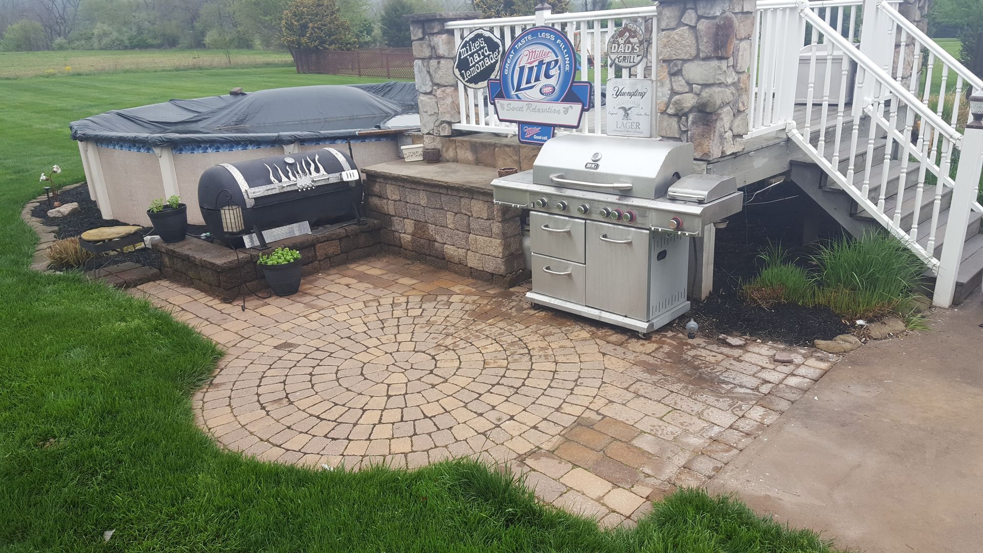 Backyard patio with grill, built-in outdoor kitchen, brick pavers, and stairs leading to a deck